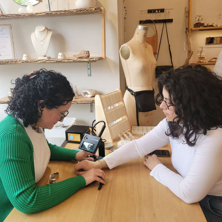 Hands-on artisan jewelry studio scene: two women at a wooden counter, one inspects a bracelet on the other's wrist with a digital loupe, necklace displays and a dress form in the background.