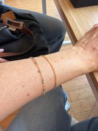Close-up of a forearm resting on a wooden cafe table with two delicate gold bracelets, leather handbag strap and gray pants visible over a wooden floor in a casual coffee shop setting