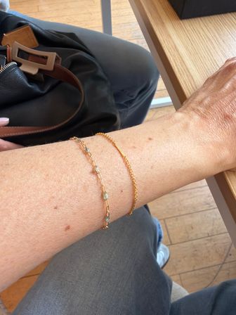 Close-up of a forearm resting on a wooden cafe table with two delicate gold bracelets, leather handbag strap and gray pants visible over a wooden floor in a casual coffee shop setting