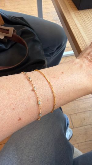 Close-up of a forearm resting on a wooden cafe table with two delicate gold bracelets, leather handbag strap and gray pants visible over a wooden floor in a casual coffee shop setting