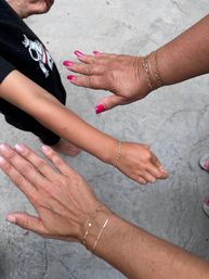 Three hands and forearms held over an outdoor concrete patio: two adults with bright pink manicures wearing delicate gold chain bracelets and a child's arm with a colorful beaded bracelet — playful family jewelry close-up.