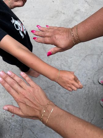 Three hands and forearms held over an outdoor concrete patio: two adults with bright pink manicures wearing delicate gold chain bracelets and a child's arm with a colorful beaded bracelet — playful family jewelry close-up.