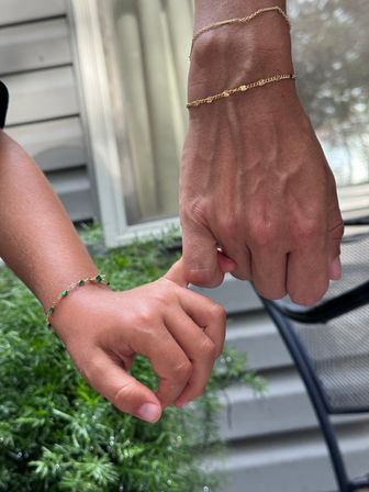 Adult and child linking pinky fingers on a house porch, gold bracelets and a green-beaded bracelet visible, window, siding and leafy backyard plants in soft focus.
