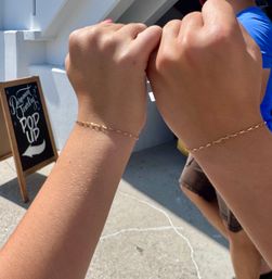 Two people bumping fists to show matching delicate gold chain bracelets outside a sidewalk pop-up on a sunny day
