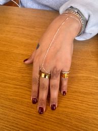 Close-up of a hand with burgundy nail polish resting on a wooden table, showcasing chunky gold rings, a delicate chain hand bracelet connecting to the wrist, layered bracelets and a small heart tattoo.