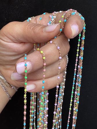 Close-up of a hand holding multiple delicate gold chains strung with tiny pastel beads (turquoise, pink, green, yellow) against a black background, colorful beaded necklace jewelry.