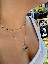 Close-up of a person wearing layered silver necklaces — black-stone eye pendant and delicate black-beaded choker — in a white tank top at an outdoor pop-up market with a chalkboard sign.