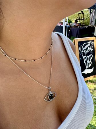 Close-up of a person wearing layered silver necklaces — black-stone eye pendant and delicate black-beaded choker — in a white tank top at an outdoor pop-up market with a chalkboard sign.