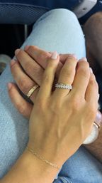 Close-up of clasped hands resting on light-blue jeans in an airplane seat — a romantic travel moment featuring a diamond eternity ring, textured gold band, delicate gold chain bracelet and wristwatch.