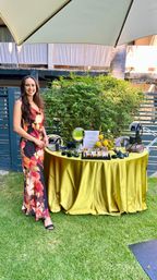 Smiling woman in a floral maxi dress stands by a round table draped in gold satin under a patio umbrella at a sunny outdoor garden setup displaying sunglasses, mirrors and small floral arrangements on green lawn.