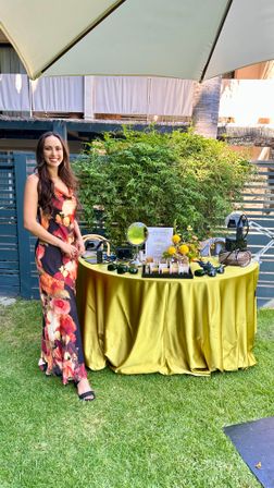 Smiling woman in a floral maxi dress stands by a round table draped in gold satin under a patio umbrella at a sunny outdoor garden setup displaying sunglasses, mirrors and small floral arrangements on green lawn.