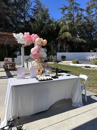 Sunlit backyard patio event with pastel pink, peach and white balloon arch beside a white-linen registration table, palm trees and outdoor seating