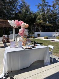 Sunny backyard patio party setup with pink, peach and white balloon arch, white-clothed welcome table and palm trees