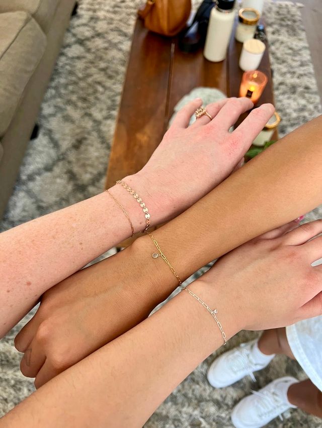 Three stacked wrists wearing delicate gold and silver chain bracelets and a ring, over a rug and wooden coffee table with candles and a water bottle in a cozy living room