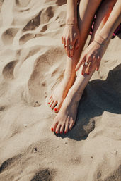 Close-up of a person's bare feet and hands with red nail polish, gold anklets and rings on a sunlit sandy beach