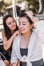 Candid outdoor portrait in a sunny park: a woman in a white tied shirt and smartwatch smiles while a friend adjusts an earring or clip behind her ear.