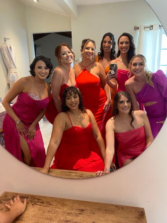 Mirror selfie of eight bridesmaids and friends in a bright bridal suite, smiling and posing in coordinated red, pink and orange formal dresses — festive getting-ready photo.