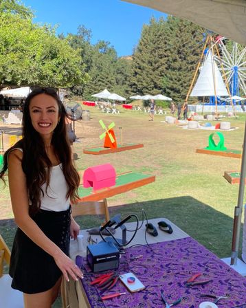 Smiling woman at a vendor table under a tent at a sunny outdoor festival with colorful mini-golf obstacles, picnic seating and a ferris wheel in a tree-lined park.