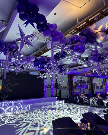 Purple-and-silver balloon garlands with oversized metallic star balloons suspended over a waterfront ballroom, purple uplighting, decorative gobo patterns projected on a white dance floor, lounge seating, and city skyline visible through floor-to-ceiling windows.
