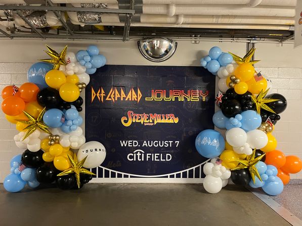 Colorful balloon arch framing a large concert poster for classic rock bands with bold logos and an event date, displayed in a stadium concourse.