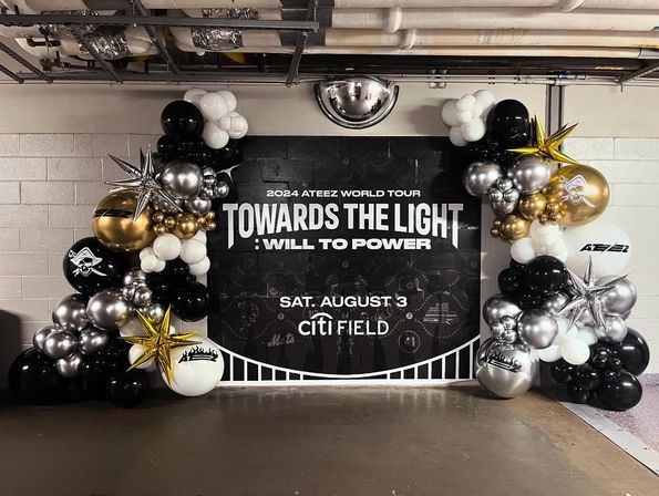 Stadium corridor display: black 'TOWARDS THE LIGHT' tour backdrop framed by festive gold, silver, black and white balloon garlands with star-shaped foil balloons.