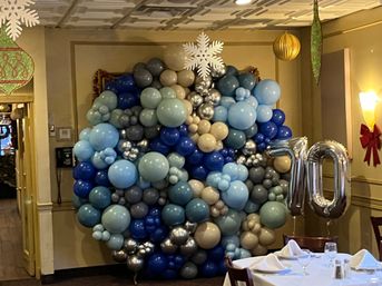 Indoor party scene with a large organic balloon wall in shades of blue, gray and metallic silver, topped by a white snowflake and flanked by silver “70” foil number balloons next to a set dining table.