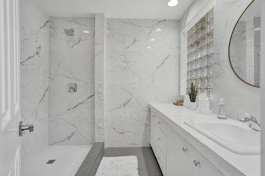Bright modern marble bathroom with walk-in shower, glass-block window, long white vanity, round mirror, potted plant and spa-like residential design