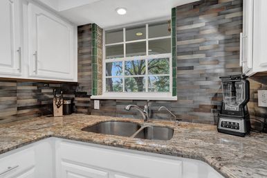 Cozy kitchen sink area with double stainless basin, chrome faucet, granite countertop, gray stacked-tile backsplash and window view of trees