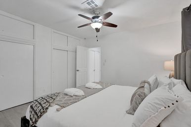 Bright minimalist bedroom interior with ceiling fan, white walls, sliding closet doors, and a large bed dressed in white linens, decorative throw and bedside lamp.