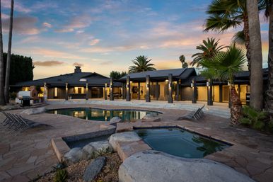 Luxury modern single-story home with lit covered patio, stone pool and hot tub on a palm-lined backyard terrace at sunset, lounge chairs and outdoor grill