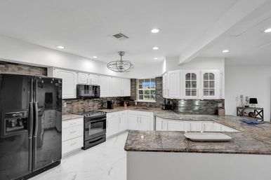 Spacious modern U-shaped kitchen with white cabinets, granite countertops and island, black appliances, glass-front uppers, mosaic tile backsplash, pendant and recessed lighting, and a window over the sink.