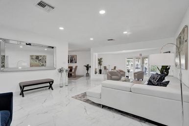 Bright modern lounge with glossy white marble floors, white sectional sofa, beige armchairs, large decorative mirror, potted plants, floor lamp, and glass doors opening to an outdoor pool area.