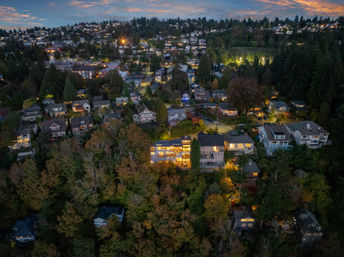Aerial drone view of a hillside residential neighborhood at dusk, cozy lit homes and multi‑story houses nestled among dense trees with winding streets and distant city lights under a colorful twilight sky