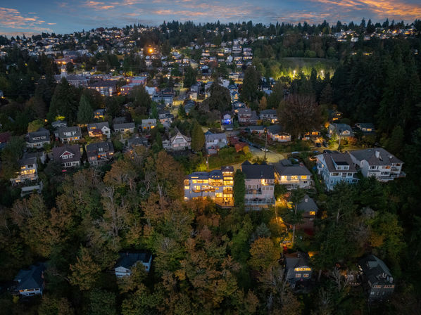 Aerial drone view of a hillside residential neighborhood at dusk, cozy lit homes and multi‑story houses nestled among dense trees with winding streets and distant city lights under a colorful twilight sky