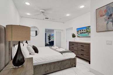 Bright, spa-like modern bedroom with king bed and upholstered headboard, wooden nightstand and lamp, wall-mounted TV showing a pool, six-drawer wood dresser, white marble-look floor, ceiling fan and minimalist wall art