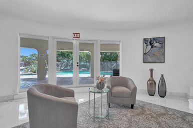 Bright modern indoor lounge with two gray armchairs and a glass side table, looking through French doors to a turquoise resort-style pool and palm-lined patio; abstract wall art and decorative vases.