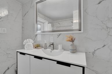Spa-like marble bathroom vanity with white countertop and integrated sink, chrome faucet, large mirror, fan-folded towel, travel-size toiletries on a tray, pump soap, and a striped vase with dried flowers.