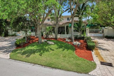 Mediterranean-style two-story house with tile roof, gated paver driveway, manicured curved lawn, red-mulched tropical landscaping and shade trees on a sunny day.