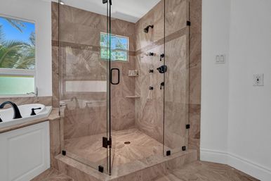 Spa-like glass-enclosed marble walk-in shower with black fixtures and built-in shelves beside a white soaking tub, bright bathroom with palm-tree window view