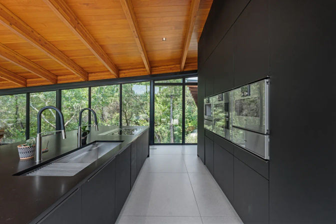 Sleek modern kitchen with matte-black island and cabinets, integrated stainless sink and faucets, floor-to-ceiling windows framing lush forest views and a warm wooden beamed ceiling.