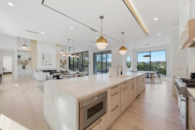 Sunlit modern open-concept kitchen and living area with a long marble island, brass globe pendant lights, light wood cabinetry with built-in microwave, stainless steel range, dining nook by large windows with treed view, and light hardwood floors.