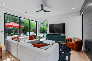 Bright modern living room with white sectional, orange accent pillows and rust velvet armchair, wall-mounted TV on teal console, black ceiling fan, large glass doors opening to a backyard patio with red umbrella and lush greenery.