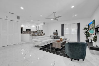 Bright modern open-concept living room and kitchen with white marble floors, white sectional sofa, bar stools, ceiling fan, wall-mounted TV showing a pool, green accent chair and large curtained window.