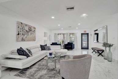 Bright modern living room with white leather sectional, navy accent chairs, glass coffee table on gray rug, marble floors, abstract wall art and large windows