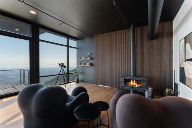 Modern mountain-view living room with floor-to-ceiling windows and balcony overlooking a valley, cozy sculptural armchairs, telescope by the glass, wood-burning stove with glowing fire, vertical wood-paneled wall and minimalist shelves.