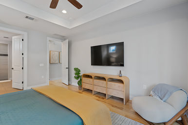 Modern bedroom with wall-mounted TV, curved wood media console, teal-and-yellow bedding, cozy accent chair, ceiling fan and open door to en-suite bathroom