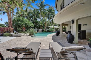 Sunny tropical backyard oasis with curved modern house, covered patio, stone pool deck, two loungers and a kidney-shaped pool framed by palm trees.