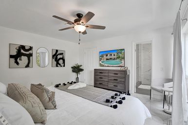 Bright, serene master bedroom with king bed dressed in textured pillows and a tasseled throw, ceiling fan, wood dresser with wall-mounted TV showing a pool, potted plant, natural light from a window, and an open doorway to a marble-tiled ensuite bathroom.
