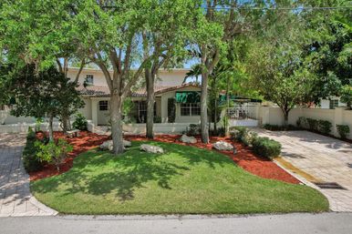 Suburban two-story stucco house with tiled roof and green awning, shaded front yard with mature trees, manicured lawn, red mulch beds, decorative rocks and paved driveways