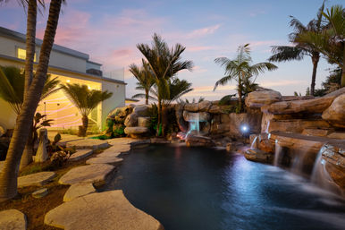 Lagoon-style backyard pool with rock waterfall, glowing LED-lit cascades, palm trees and flagstone decking beside a modern house at sunset.